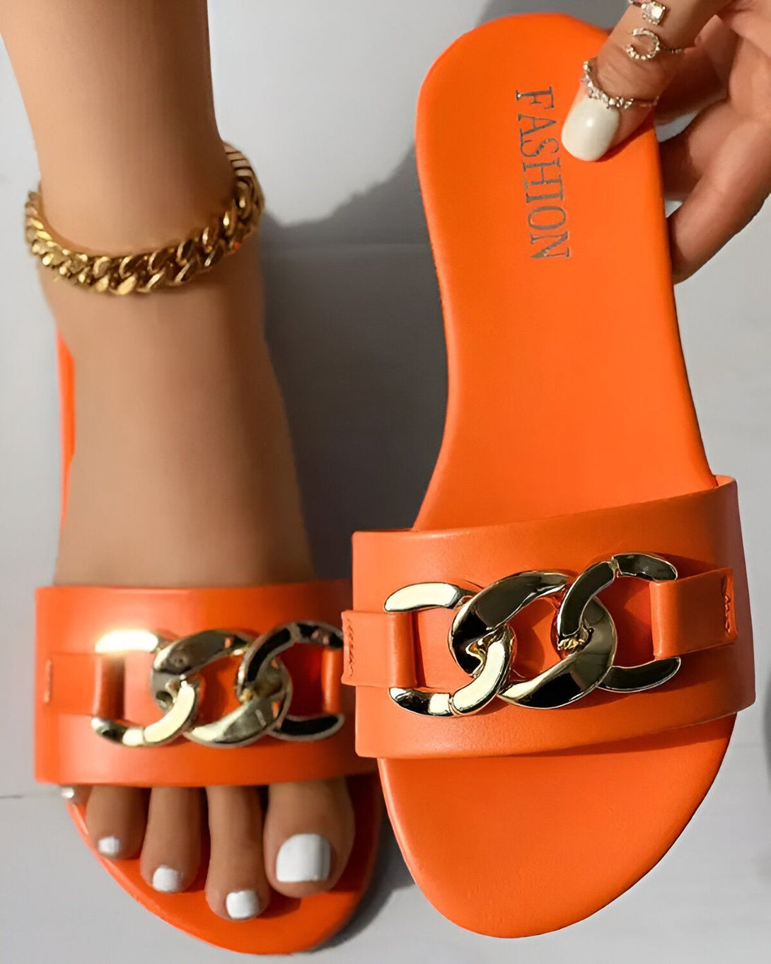 Une femme porte des sandales orange avec des chaînes dorées. Les sandales sont en plastique et ont une semelle plate. La femme a les ongles des pieds peints en blanc et porte un bracelet de cheville doré.