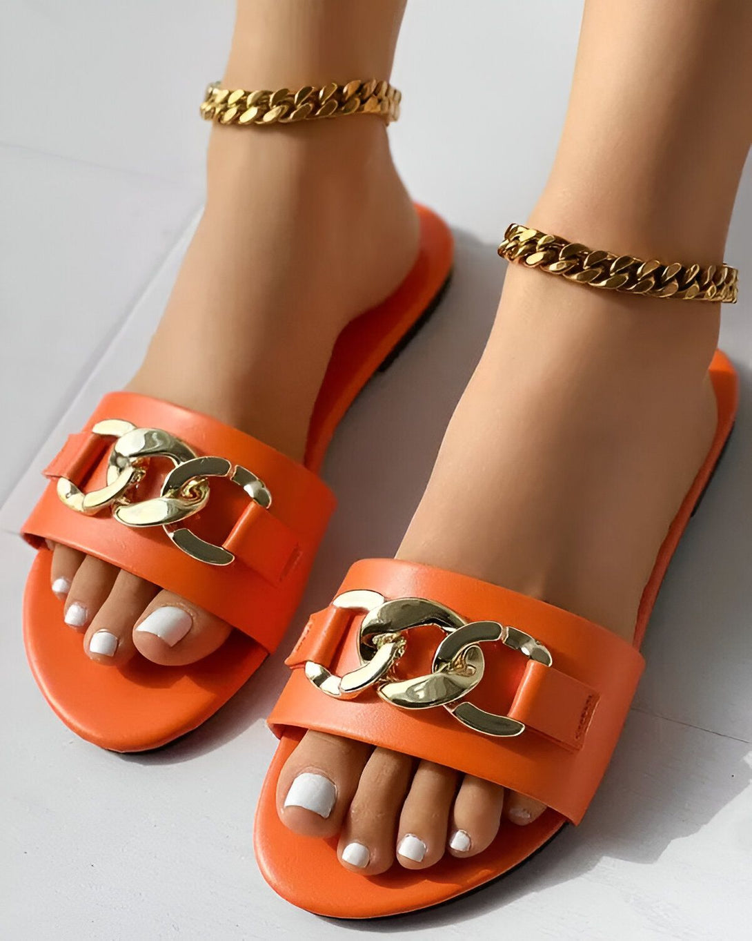 Une paire de sandales orange avec des chaînes dorées sur le dessus. Les sandales sont portées par une femme avec un bracelet de cheville doré.