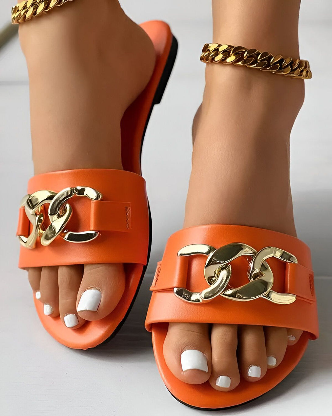 Une paire de sandales orange avec une chaîne dorée sur le dessus. Les sandales sont portées par une femme avec une peau bronzée et des ongles blancs.