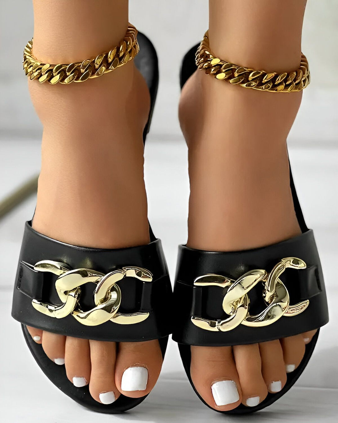 Une paire de sandales noires à talons hauts avec des chaînes dorées sur le dessus. Les sandales sont portées par une femme avec une peau bronzée et des ongles blancs.
