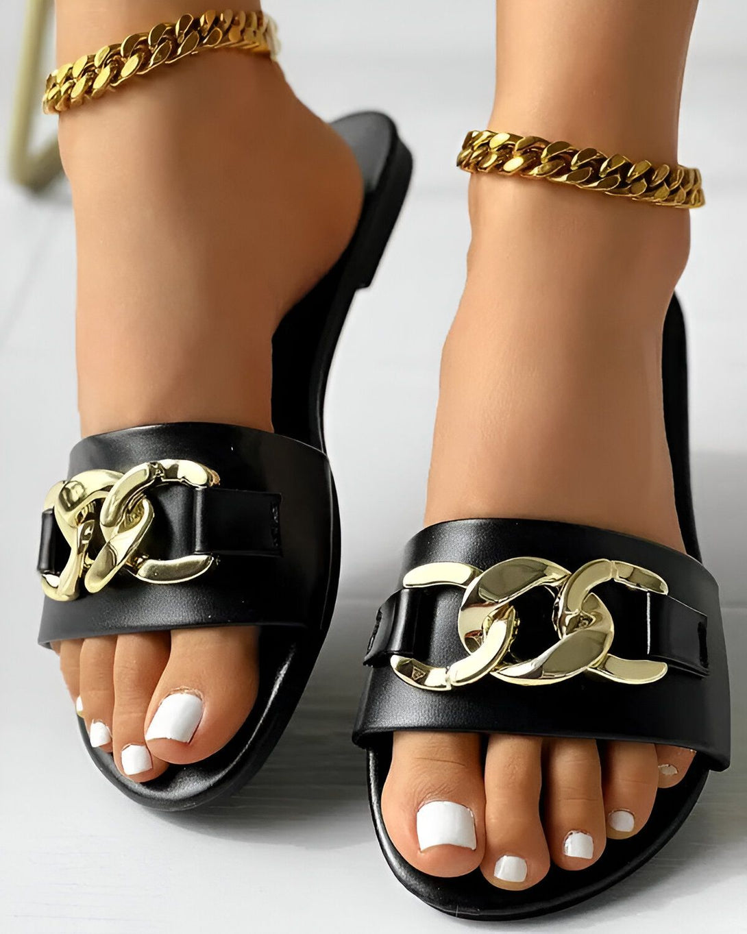 Une paire de sandales noires avec des chaînes dorées sur le dessus. Les sandales sont portées par une femme qui a les ongles des pieds peints en blanc.