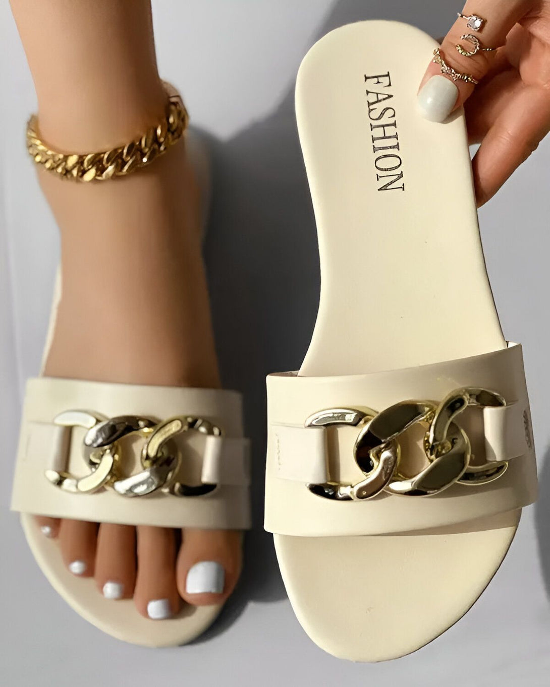 Une paire de sandales blanches avec des chaînes dorées sur le dessus. Les sandales sont portées par une femme qui a les ongles des pieds peints en blanc. La femme porte également un bracelet de cheville doré.