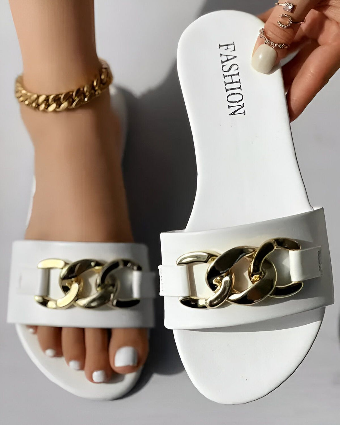 Une paire de sandales blanches avec des chaînes dorées sur le dessus. Les sandales sont portées par une femme qui a une peau bronzée et des ongles blancs. La femme porte également un bracelet de cheville doré.