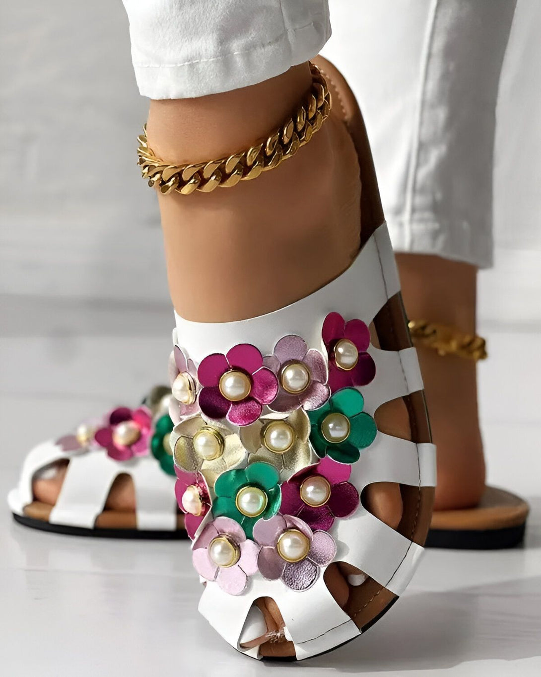 Une femme porte des sandales blanches avec des fleurs colorées et des perles. Les sandales sont en cuir et ont une semelle plate. La femme porte également un jean blanc et un chemisier blanc. Elle a les cheveux longs et noirs et porte un collier doré.