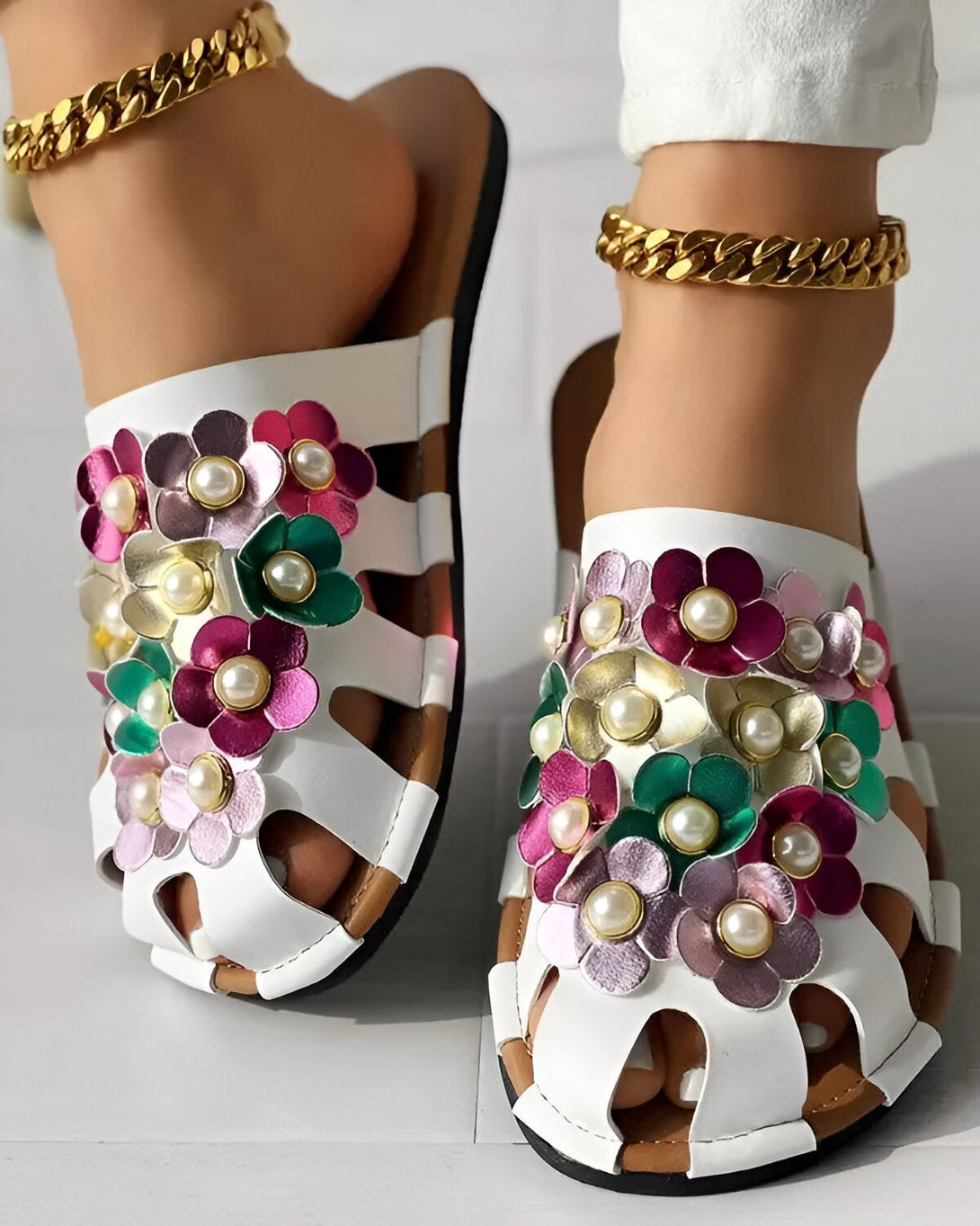 Une paire de sandales blanches à talons compensés avec des fleurs en cuir colorées et des perles. Les sandales sont portées par une femme qui a les chevilles croisées.