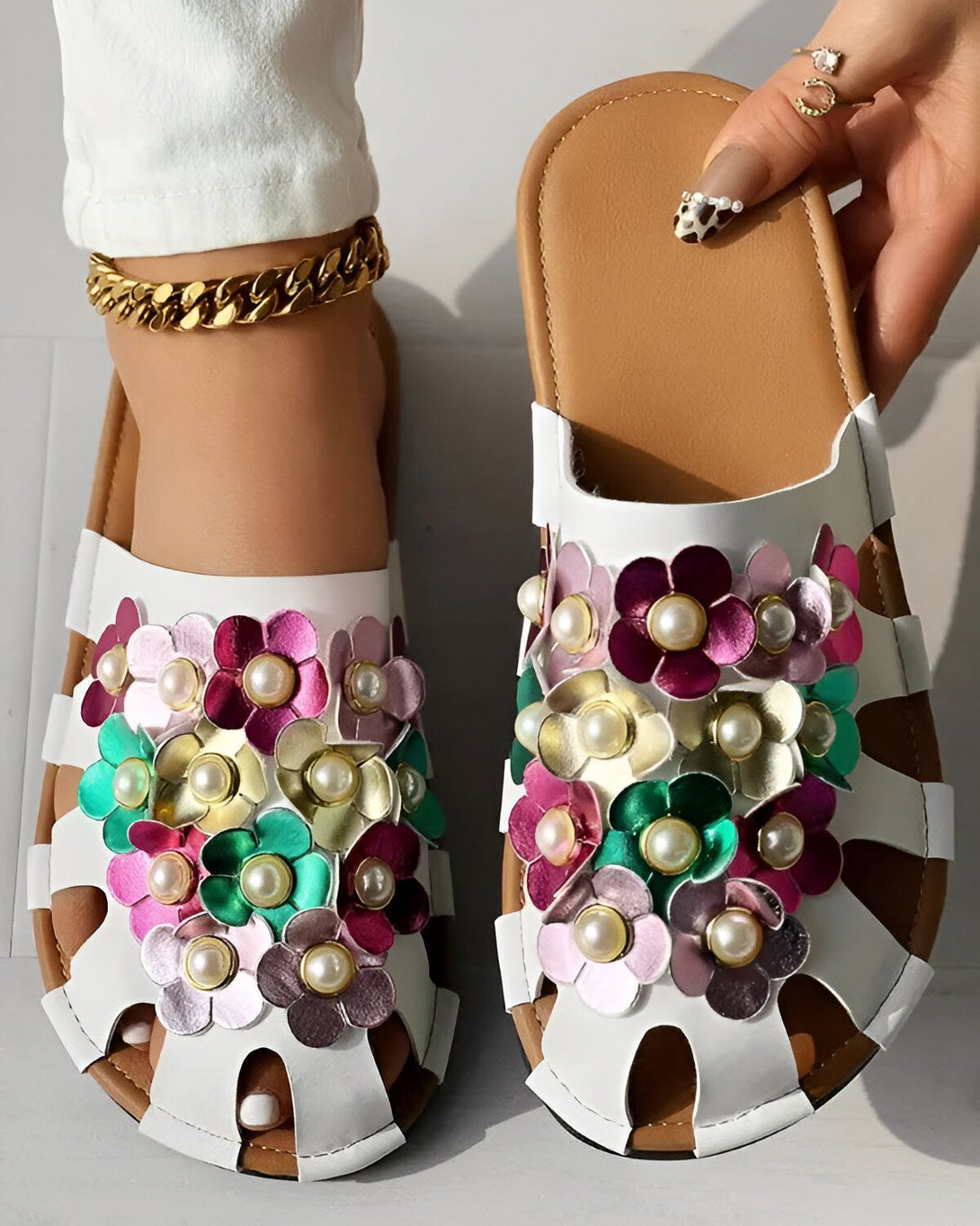 Une paire de mules blanches à fleurs colorées. Les fleurs sont en métal et en perles. Les mules sont en cuir.