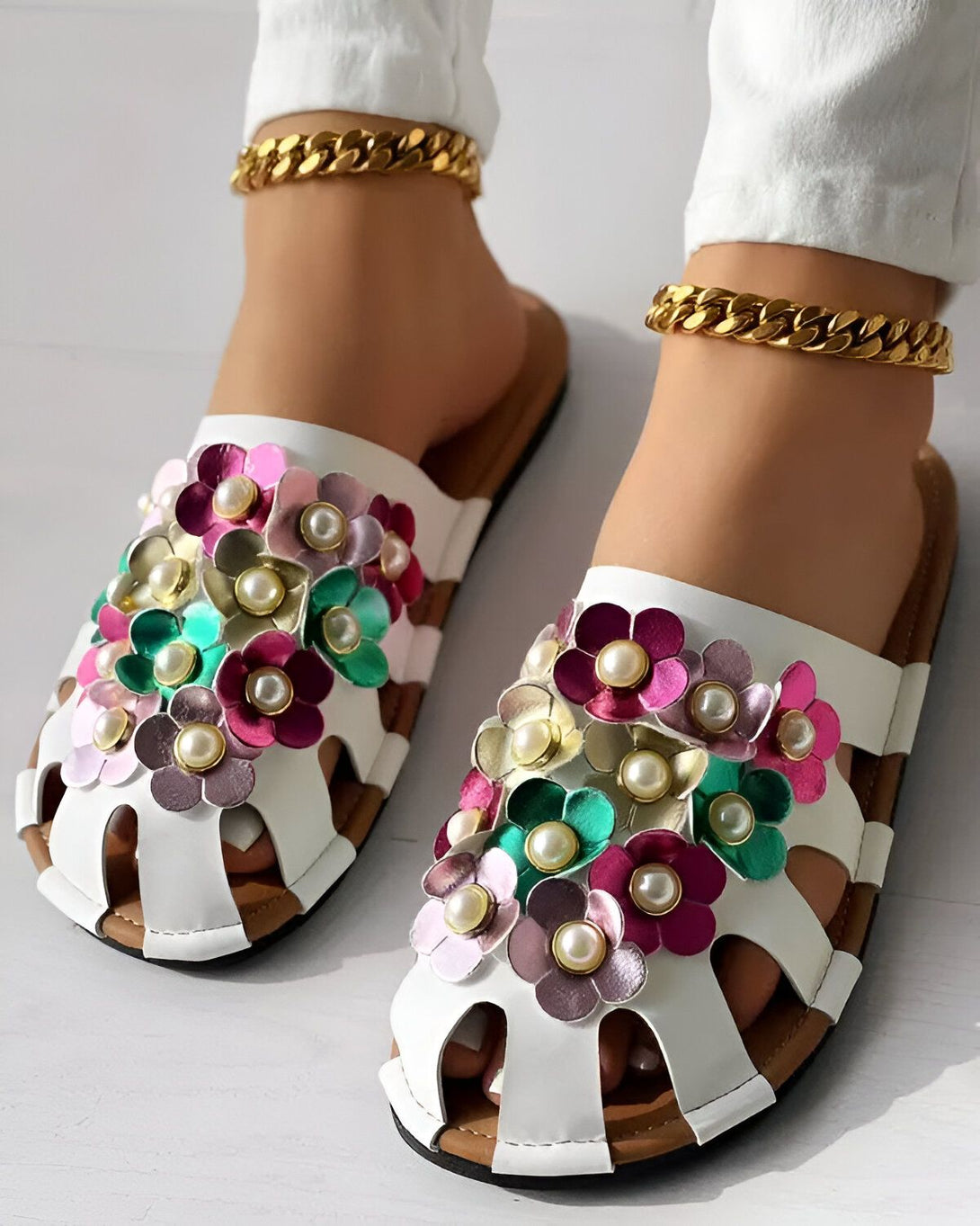 Une paire de sandales blanches à talons plats avec des fleurs en cuir colorées et des perles. Les sandales sont portées par une femme qui porte un jean blanc et des chevilles dorées.