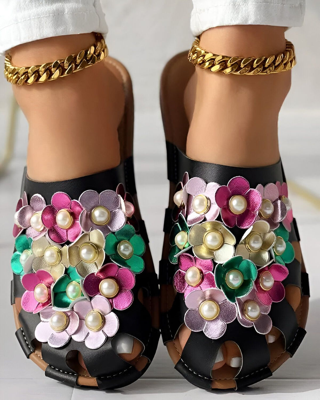 Une paire de mules noires à talons compensés, avec des fleurs en cuir colorées et des perles blanches. Les fleurs sont roses, vertes et violettes. Les mules sont portées par une femme qui a les chevilles fines et qui porte un pantalon blanc et un haut blanc. La femme est debout sur un sol en marbre blanc.