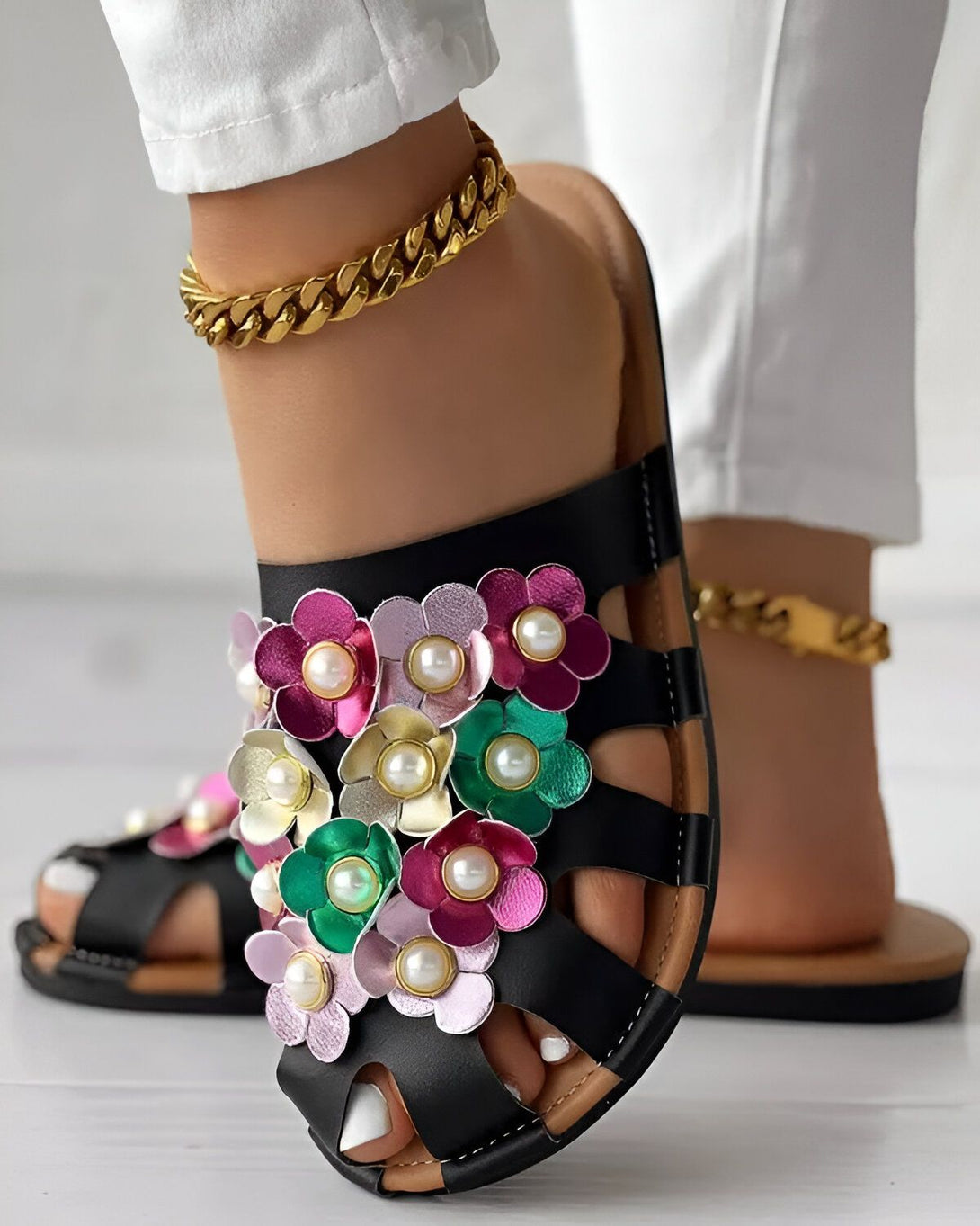 Une femme porte des sandales noires à talons compensés. Les sandales sont ornées de fleurs colorées en 3D. La femme porte également un pantalon blanc et un chemisier blanc. Elle a les cheveux longs et noirs. Elle est debout sur un sol en carrelage beige.