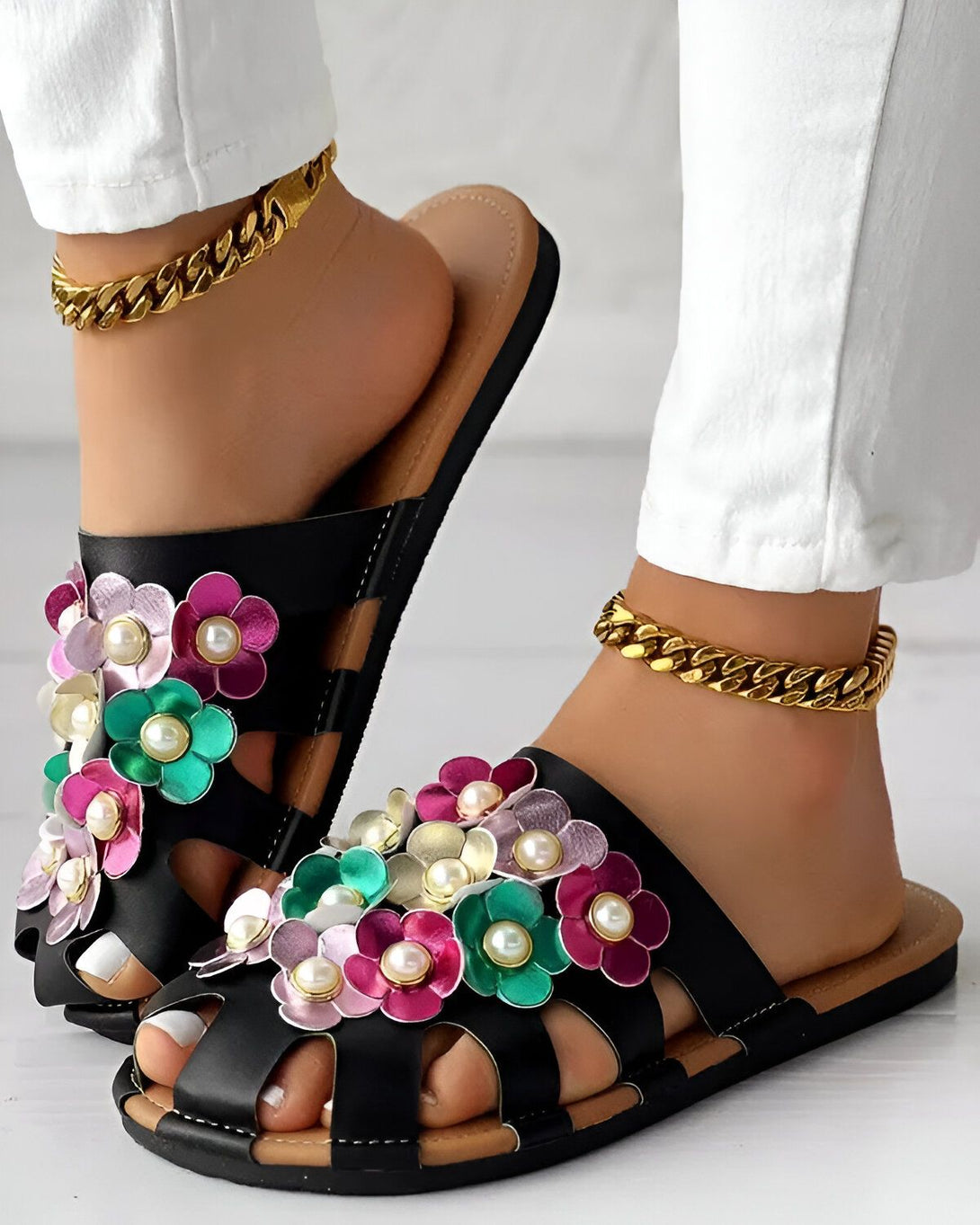 Une paire de sandales plates noires à fleurs colorées pour femmes. Les fleurs sont roses, vertes et blanches. Les sandales sont portées par une femme qui porte un jean blanc et des chevilles dorées.