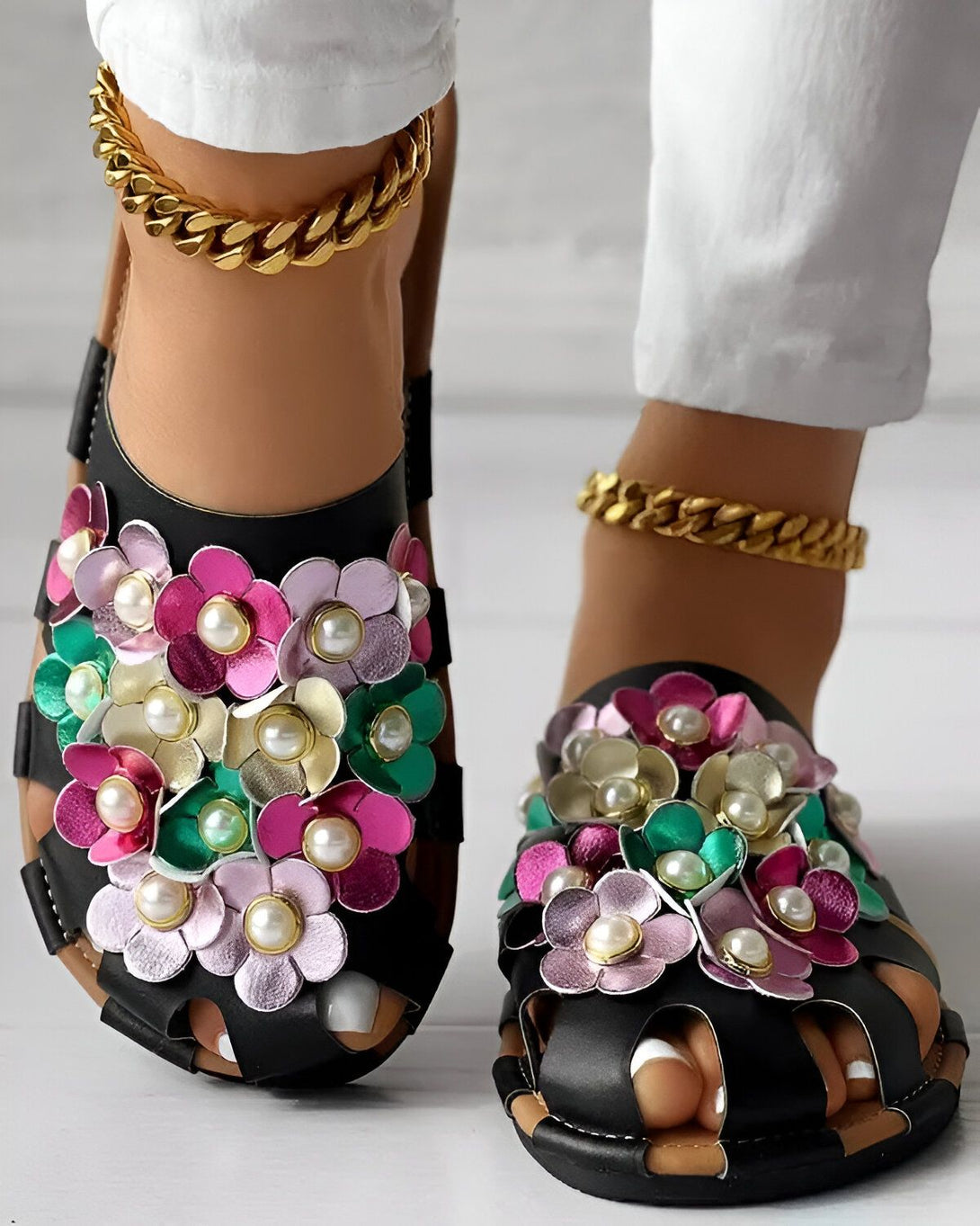 Une paire de sandales plates noires à fleurs colorées et perles blanches. Les sandales sont portées par une femme qui porte un pantalon blanc et des chevilles dorées.