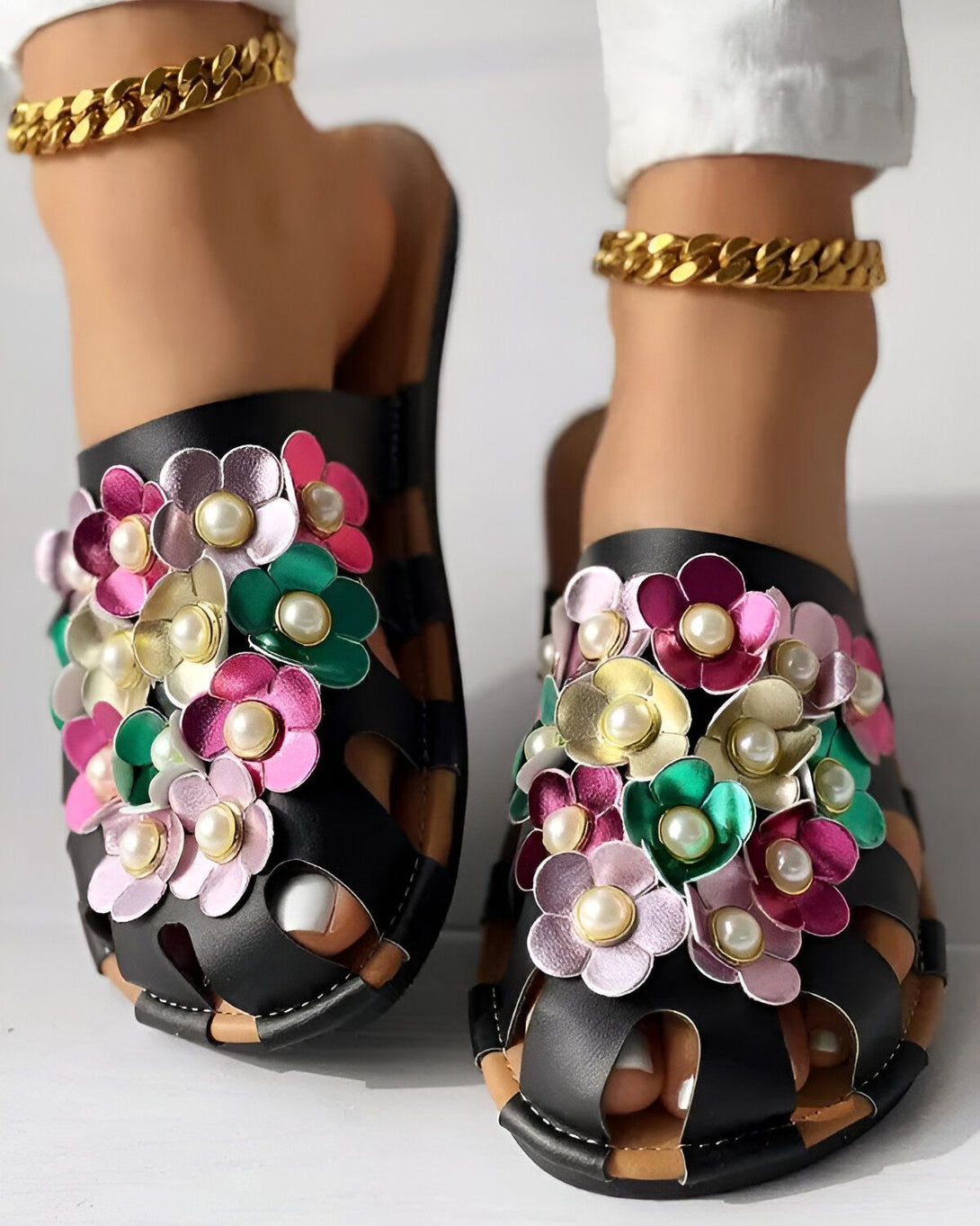 Une paire de sandales plates noires avec des fleurs en cuir colorées et des perles blanches. Les sandales sont portées par une femme avec un pantalon blanc et des chevilles dorées.