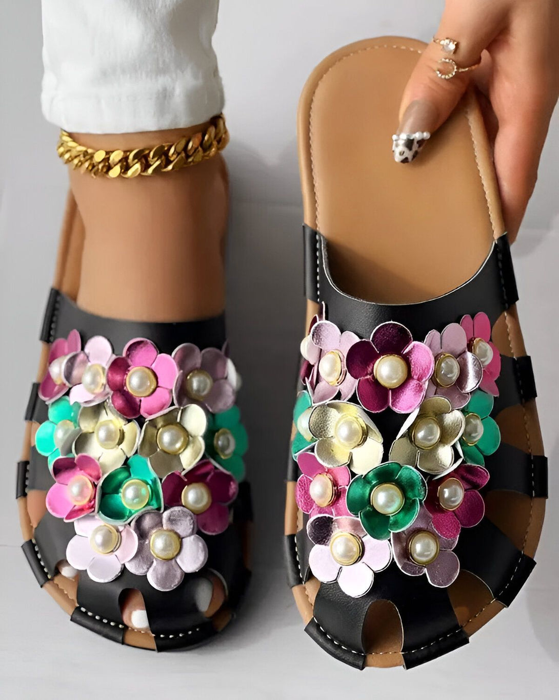 Une paire de mules plates noires à fleurs colorées. Les fleurs sont en cuir et en métal et sont de couleur rose, jaune, verte et violette. Les mules sont enfilées sur un pied droit et un pied gauche.