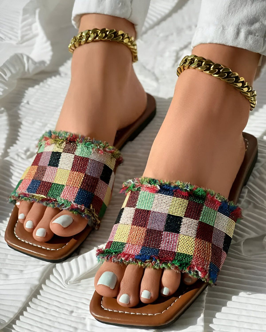 Une femme porte des sandales plates à enfiler avec un motif à carreaux. Les sandales sont marron clair et le motif est composé de carrés de différentes couleurs, notamment du rouge, du jaune, du vert, du bleu et du noir. Les sandales ont également une frange sur le devant. La femme porte également un pantalon blanc et un bracelet de cheville doré.