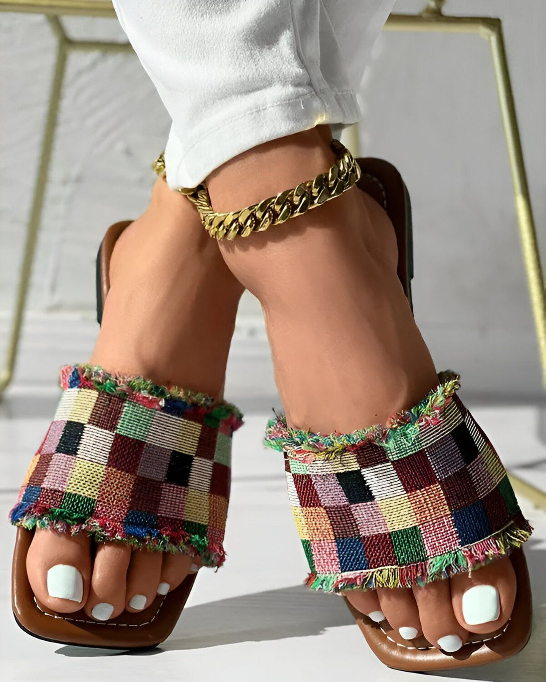 Une femme porte des sandales plates à enfiler avec une empeigne en tissu à carreaux multicolores et une semelle marron. Elle porte également un pantalon blanc et un bracelet de cheville en chaîne doré. Ses ongles sont peints en blanc.