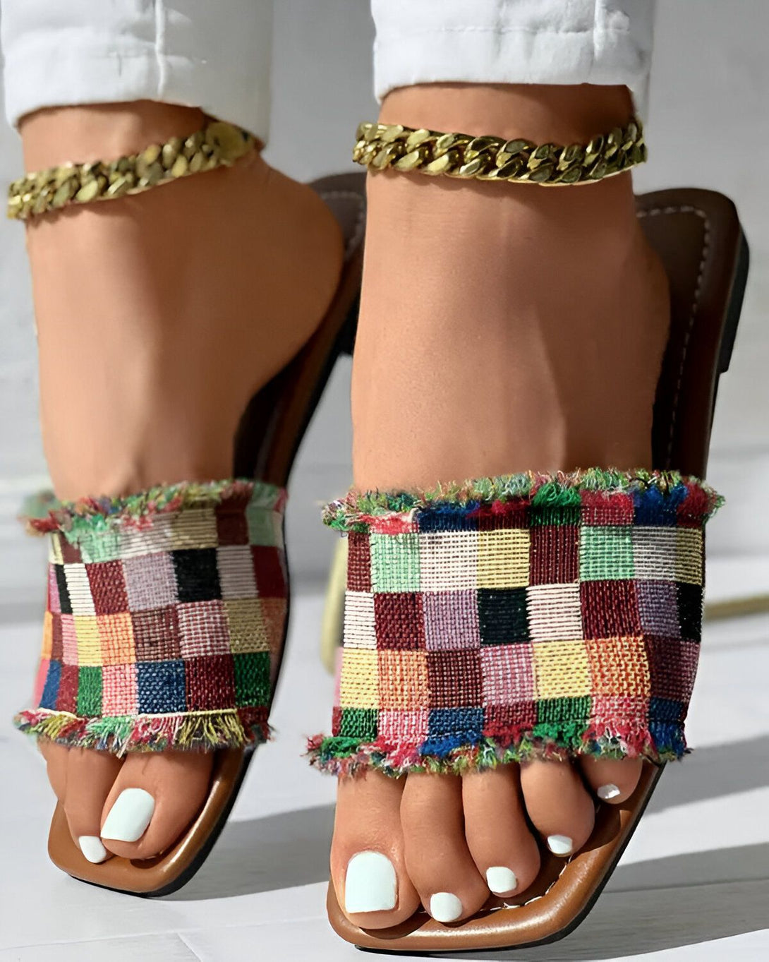 Une femme porte des sandales plates à enfiler avec une bride large et colorée. Les sandales sont faites de tissu et ont une semelle en caoutchouc. La femme porte également un pantalon blanc et un chemisier blanc. Elle a les cheveux longs et noirs et les ongles des pieds blancs.