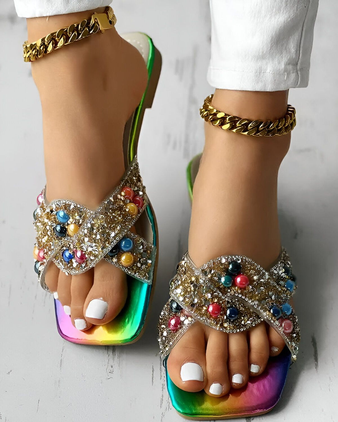 Une femme porte des sandales plates à enfiler avec des perles colorées et des détails scintillants. Les sandales ont une semelle intérieure rembourrée et une semelle extérieure antidérapante. La femme porte également un jean blanc et un bracelet de cheville doré.