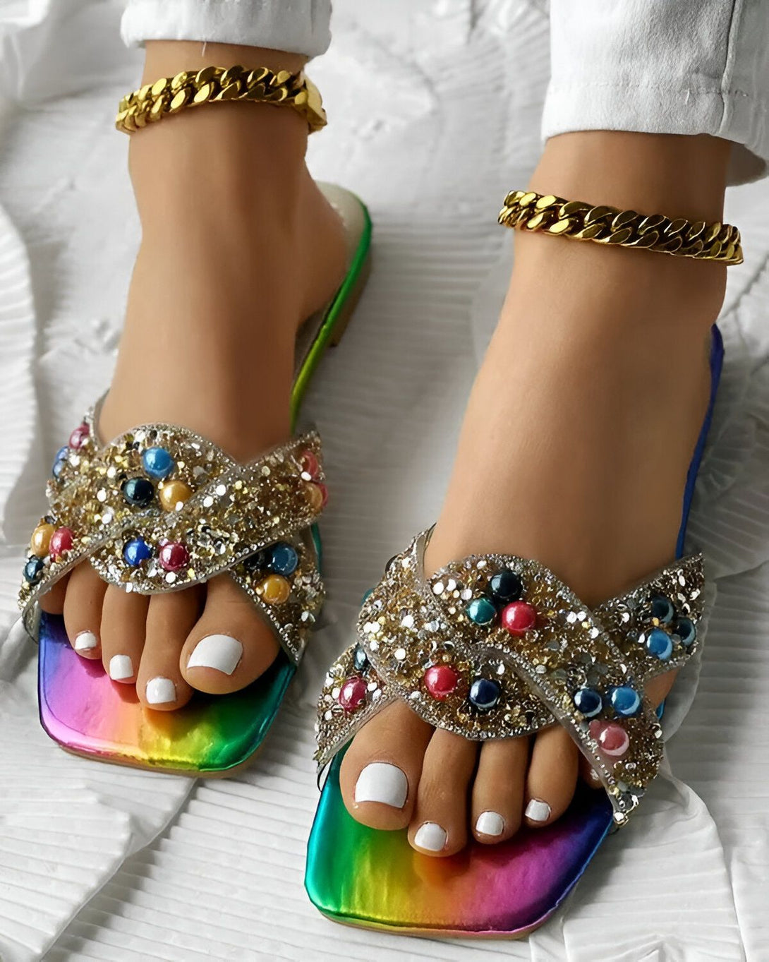 Une paire de sandales plates arc-en-ciel à bout carré avec des brides croisées et décorées de perles colorées et de paillettes. Les sandales sont portées par une femme qui a les ongles des pieds peints en blanc et qui porte des chaînes dorées aux chevilles.