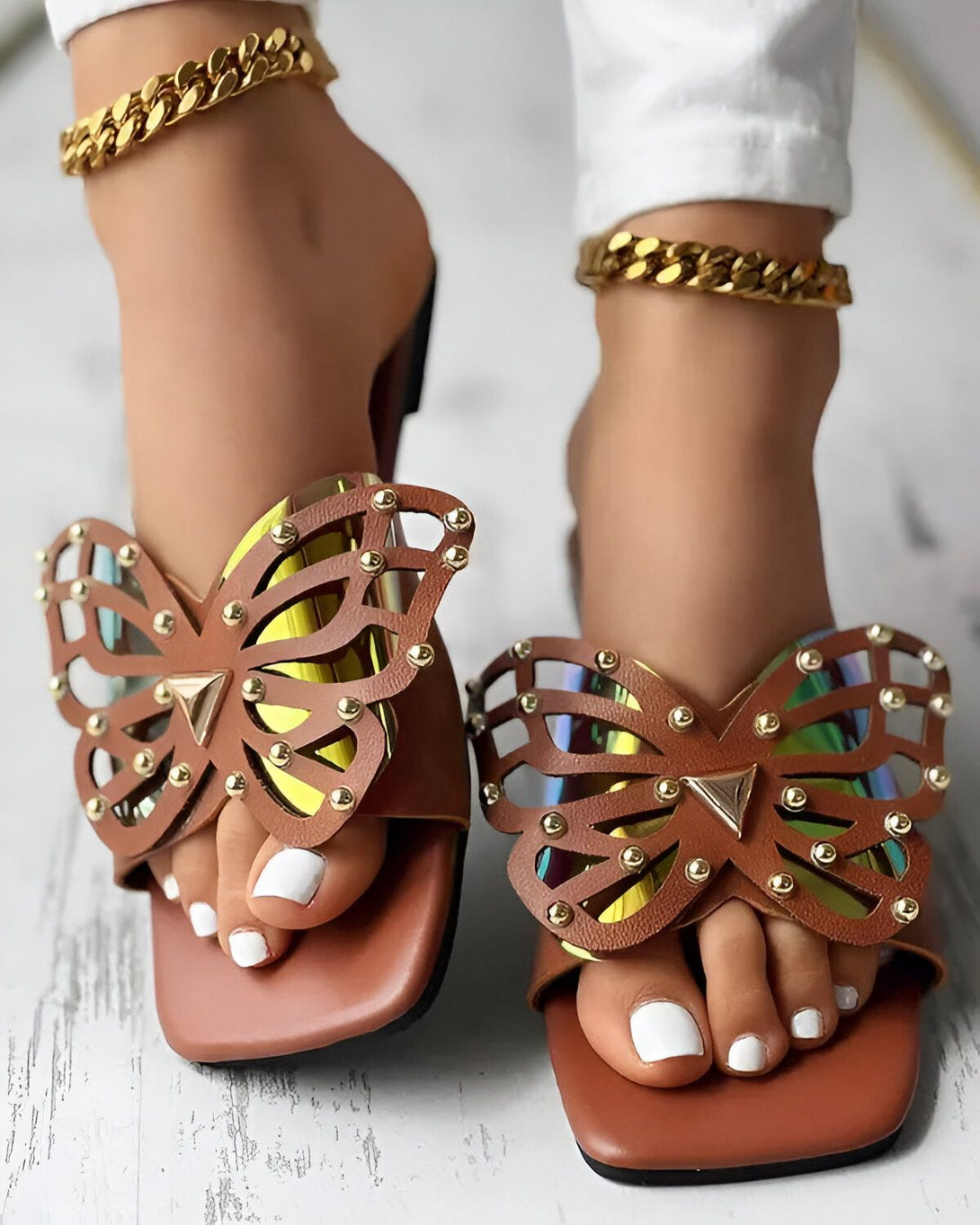 Une paire de sandales marron avec un talon carré et un bout ouvert. Les sandales sont ornées d'un papillon en métal doré.