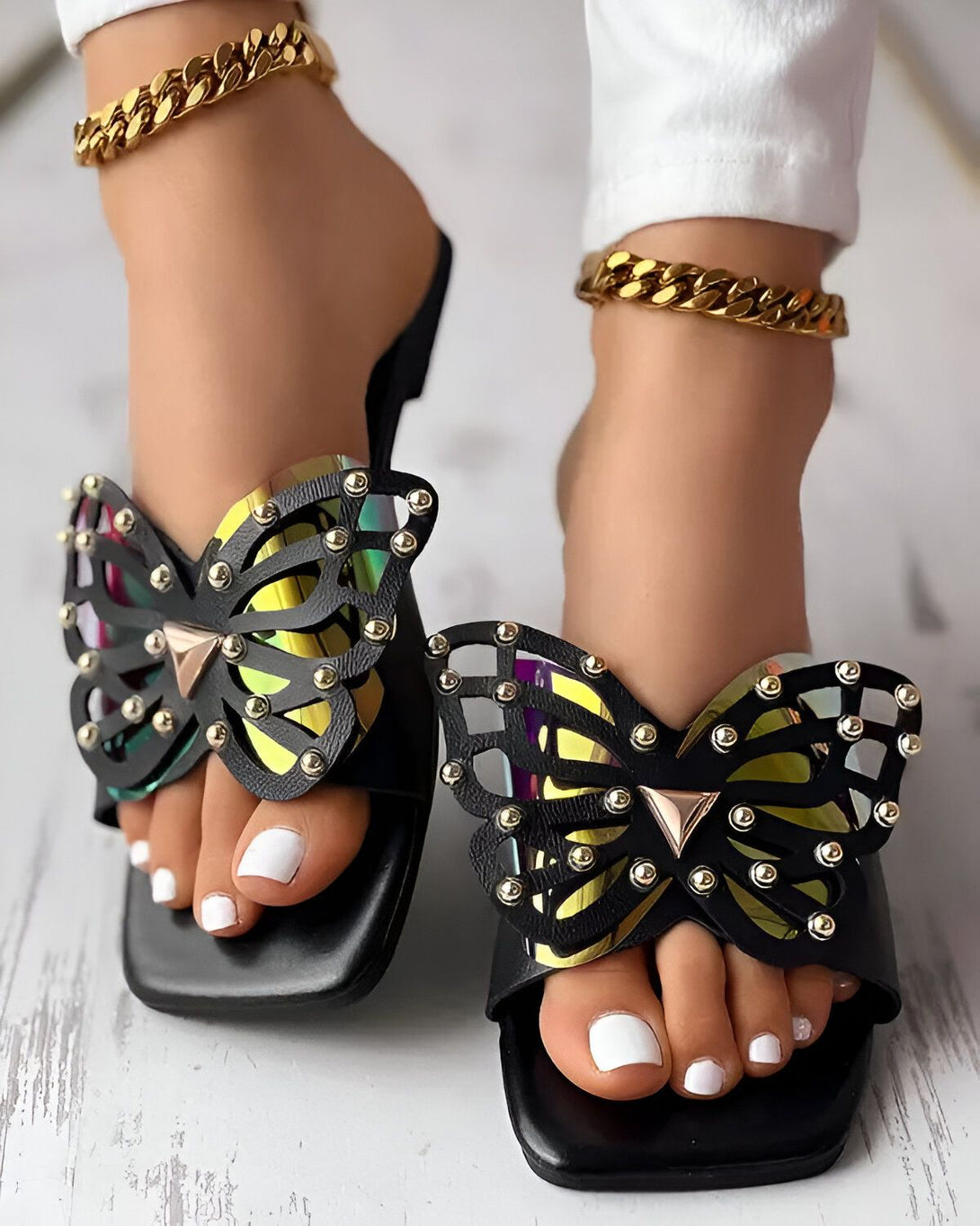 Une femme porte des sandales noires à talons avec un motif de papillon. Les sandales sont ornées de clous dorés et les ailes du papillon sont irisées.