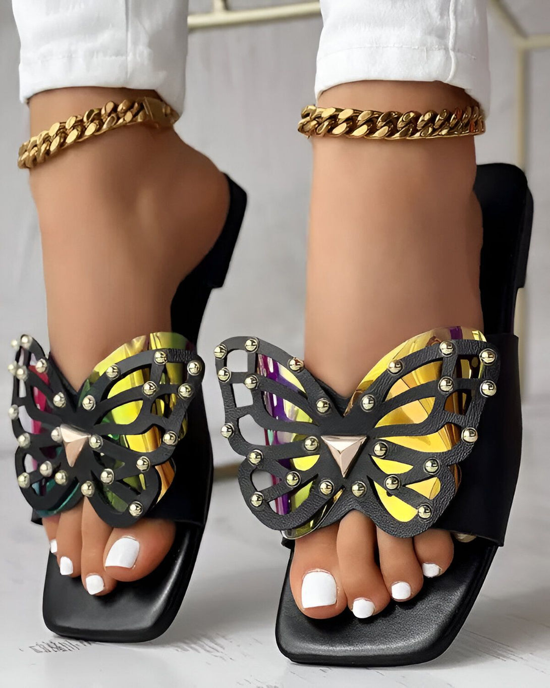 Une femme porte des sandales noires à talons hauts avec un motif de papillon doré. Les sandales sont ouvertes au niveau des orteils et ont une bride qui passe sur le dessus du pied. La femme a les ongles des pieds peints en blanc.