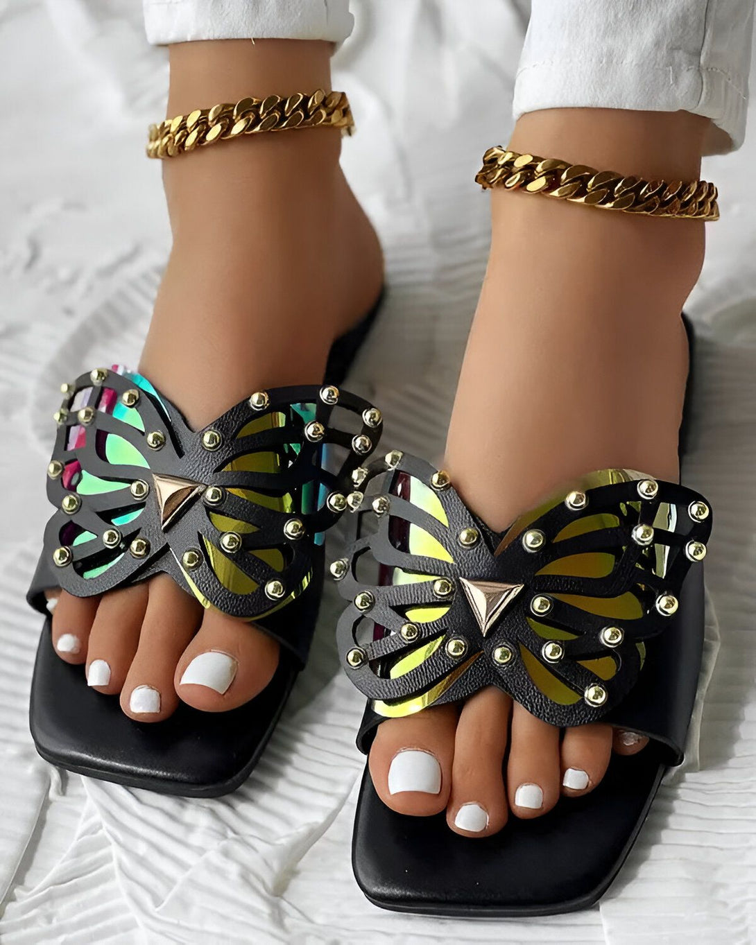 Une paire de sandales noires à talons avec un motif de papillon sur le dessus. Les sandales sont portées par une femme qui a les ongles des pieds peints en blanc.