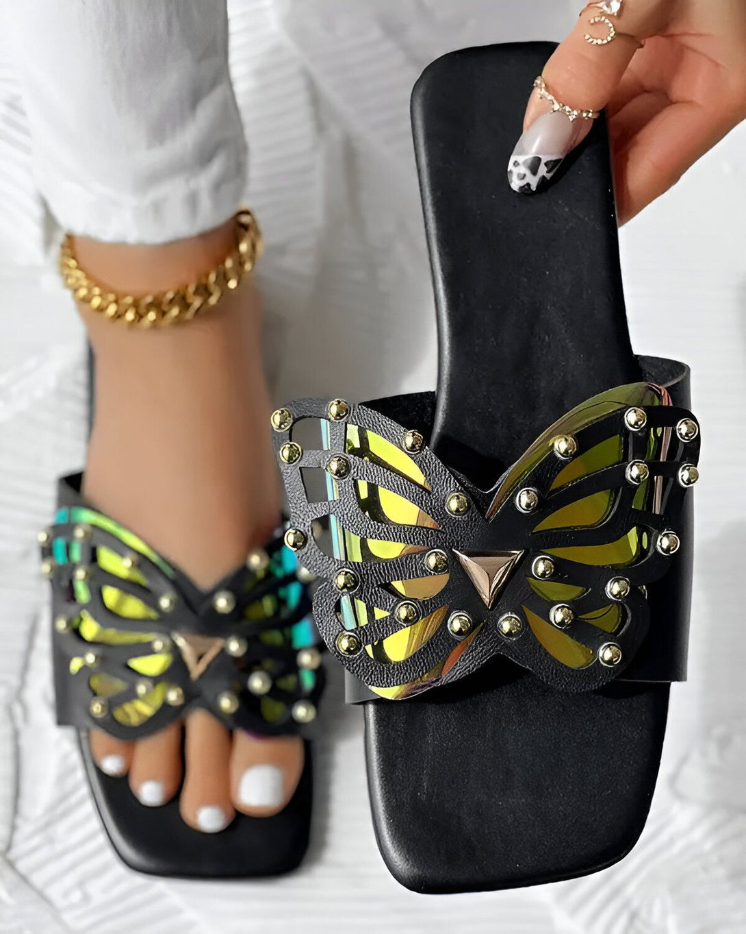 Une femme porte des sandales noires avec un papillon doré. Les sandales sont plates et ont une bride qui passe entre les orteils. Le papillon est ajouré et est décoré de petites pierres précieuses. La femme a les ongles peints en noir et blanc et porte un bracelet de cheville en or.