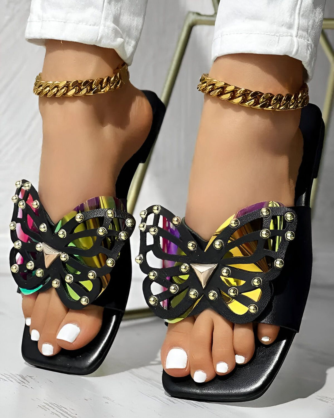 Une femme porte des sandales noires à talons hauts. Les sandales sont ornées de papillons en métal. La femme a les ongles des pieds peints en blanc. Elle porte également des bracelets de cheville en or.