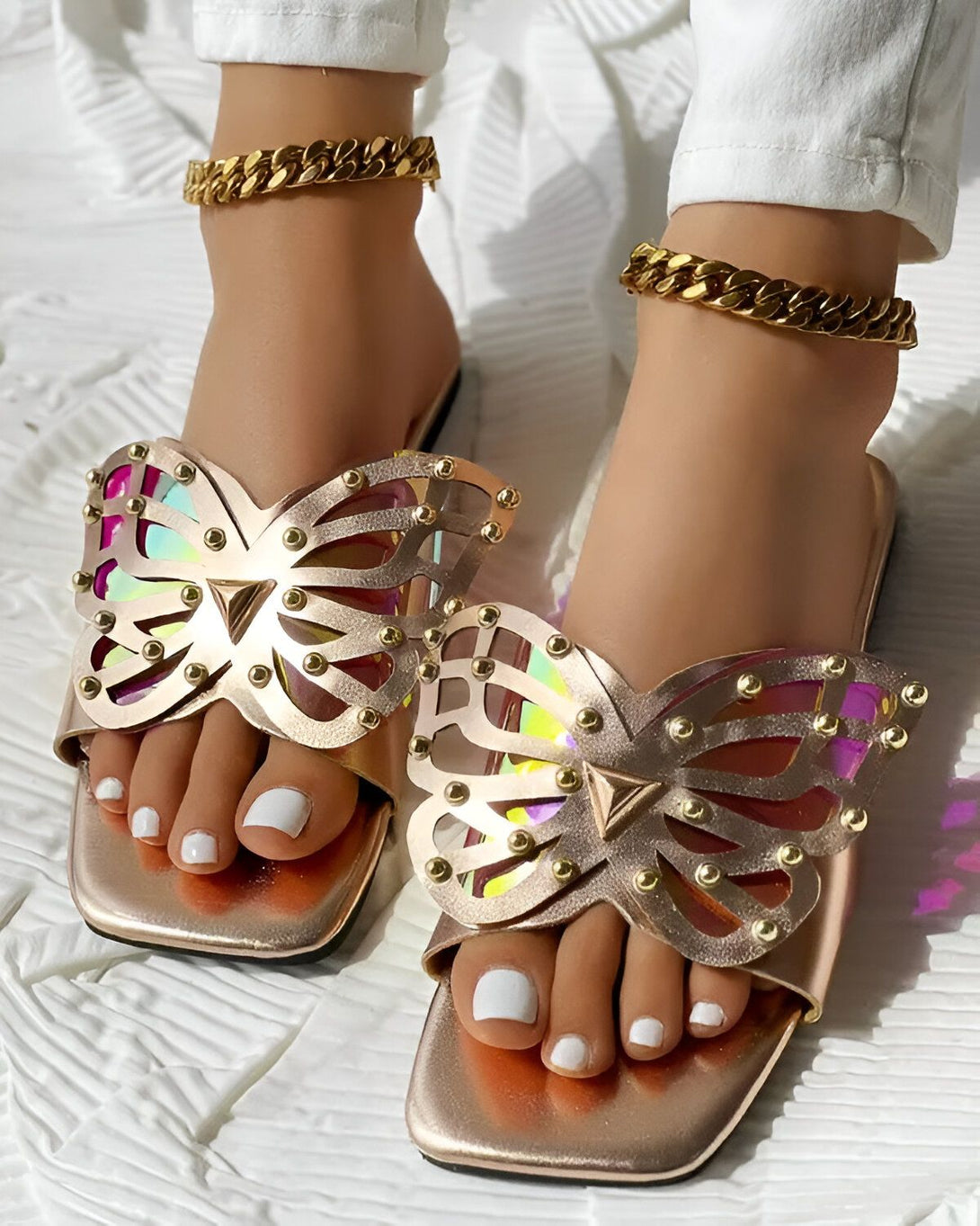 Une paire de sandales plates dorées en forme de papillon avec des détails cloutés et une semelle intérieure blanche. Les sandales sont portées par une femme avec un pantalon blanc et des bijoux dorés.