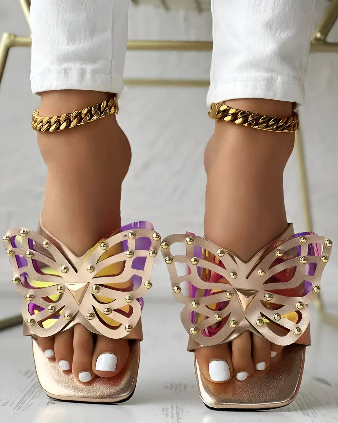 Une femme porte des sandales à talons hauts dorées en forme de papillon. Les sandales sont ornées de strass et de perles. La femme porte également un pantalon blanc et un haut blanc. Elle est assise sur une chaise et ses pieds sont posés sur le sol.