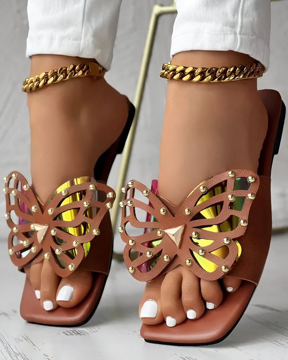 Une paire de sandales marron clair à talons carrés avec un motif de papillon doré. Les sandales sont portées par une femme avec un pantalon blanc et des bijoux en or.