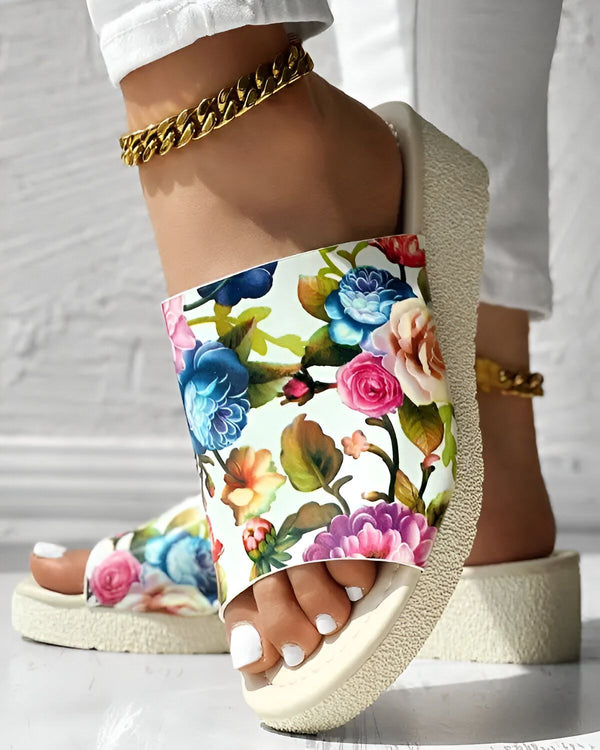 Une femme porte des tongs à fleurs blanches. Les tongs ont une semelle compensée et sont ornées d'un motif floral coloré. La femme porte également un pantalon blanc et un bracelet de cheville doré.
