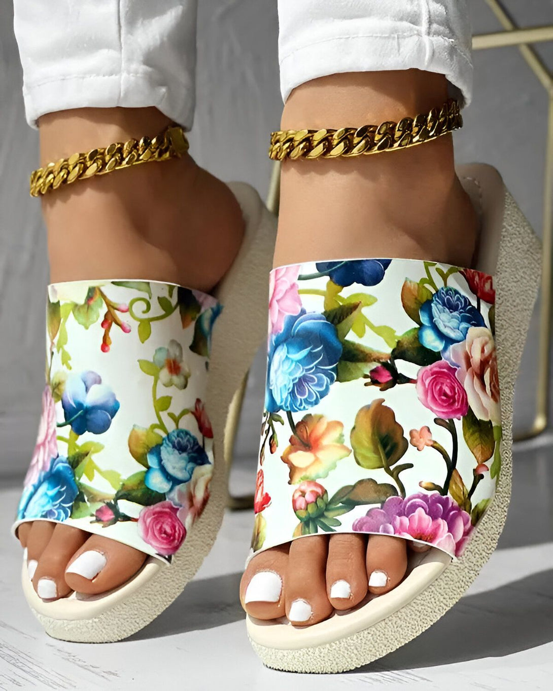 Une femme porte des sandales à fleurs avec des chaînes en or aux chevilles. Les sandales sont blanches avec des fleurs colorées. La femme porte un jean blanc et les ongles des pieds peints en blanc.