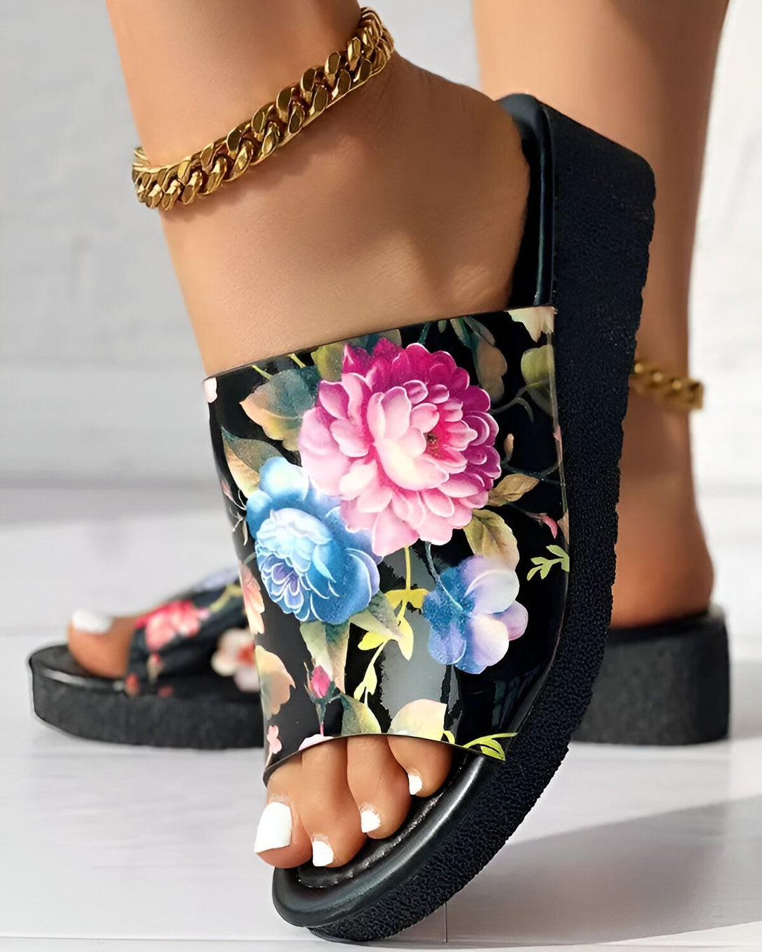 Une femme porte des sandales à plateforme noires à motif floral. Les sandales sont dotées d'une semelle épaisse et d'une bride large. La femme porte également une chaîne en or à la cheville.