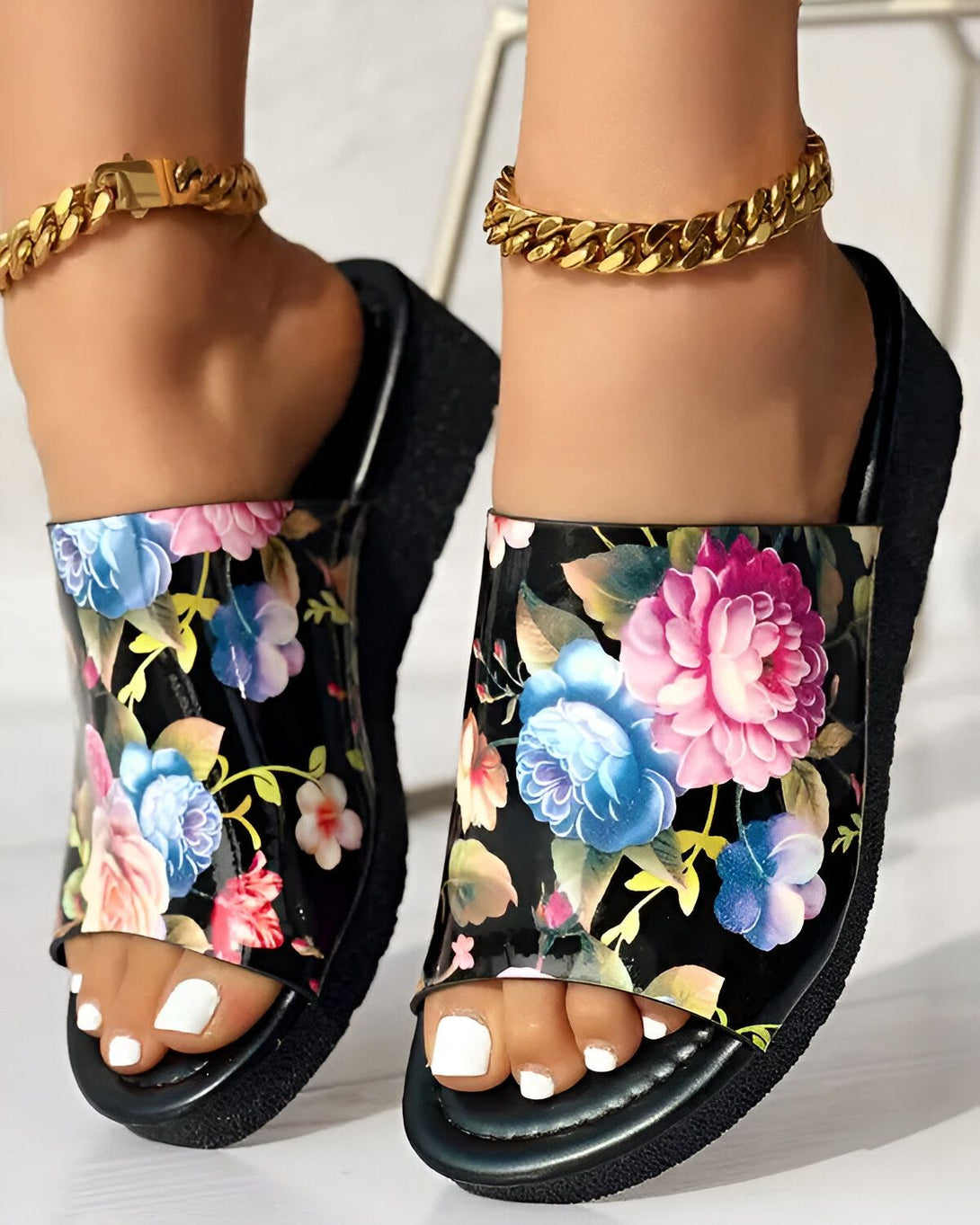 Une femme porte des sandales à talons compensés à motif floral. Les sandales sont noires avec des fleurs roses, bleues et violettes. La femme a les ongles des pieds peints en blanc et porte des bracelets de cheville en or.