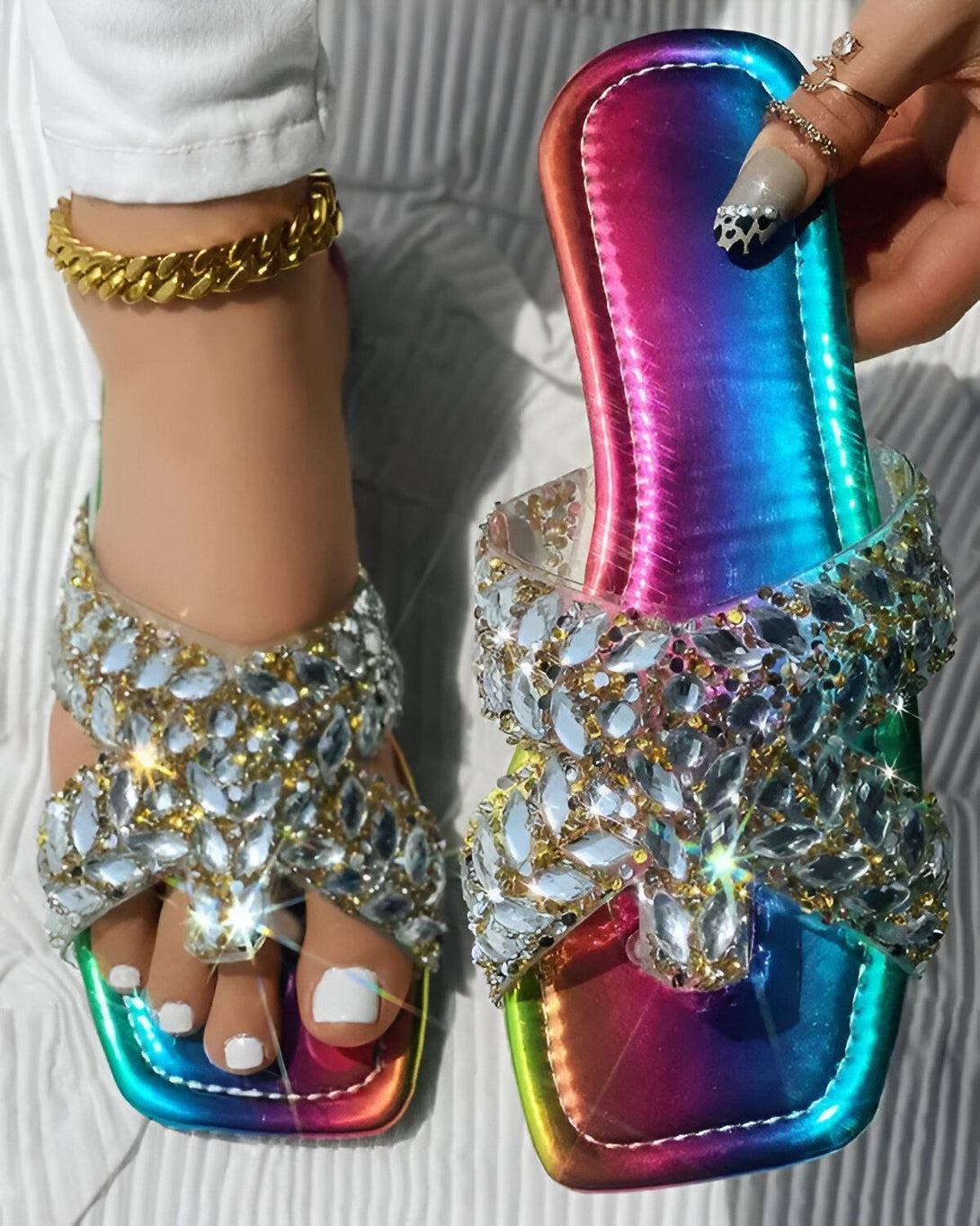 Une femme porte des sandales arc-en-ciel à talons hauts avec des cristaux. Les sandales sont faites de lanières de plastique transparentes et de cristaux de différentes couleurs. La femme a les ongles peints en noir et porte un bracelet de cheville en or.