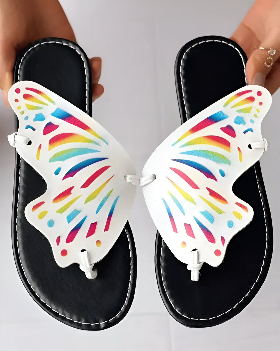 Une paire de tongs blanches en forme de papillon avec des ailes arc-en-ciel. Les tongs sont maintenues par une lanière noire qui passe entre les orteils.