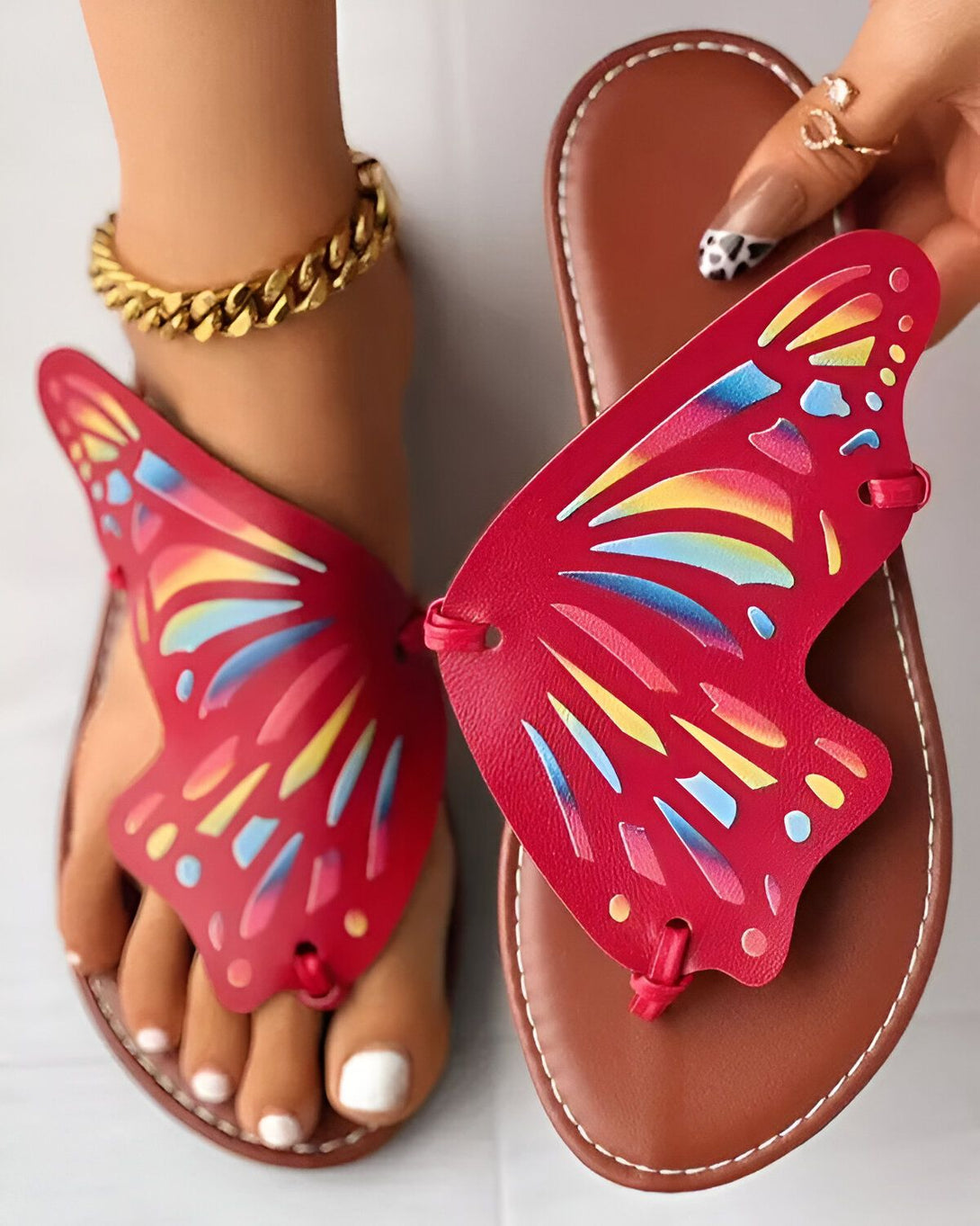 Une femme porte des sandales rouges en forme de papillon. Les sandales sont ornées d'un motif de papillons multicolores. La femme a les ongles des pieds peints en rose et porte un bracelet de cheville doré.