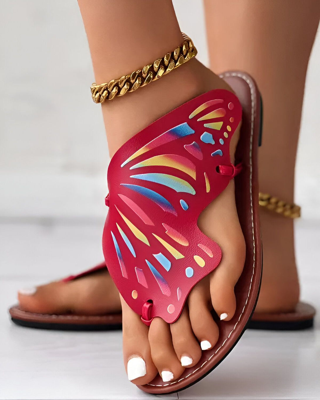 Une femme porte des sandales rouges avec un motif de papillon. Les sandales sont en cuir et ont une semelle plate. La femme a les ongles des pieds peints en blanc et porte un bracelet de cheville en or.