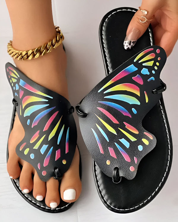 Une paire de sandales plates noires avec un motif de papillon coloré sur le dessus. Les sandales sont portées par une femme qui a une chaîne de cheville dorée et un vernis à ongles noir et blanc.