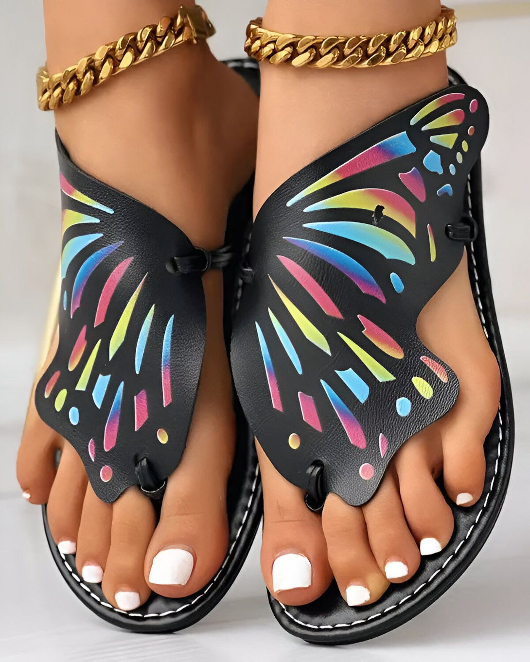 Une paire de sandales plates noires à l'effigie d'un papillon multicolore sur le dessus du pied.