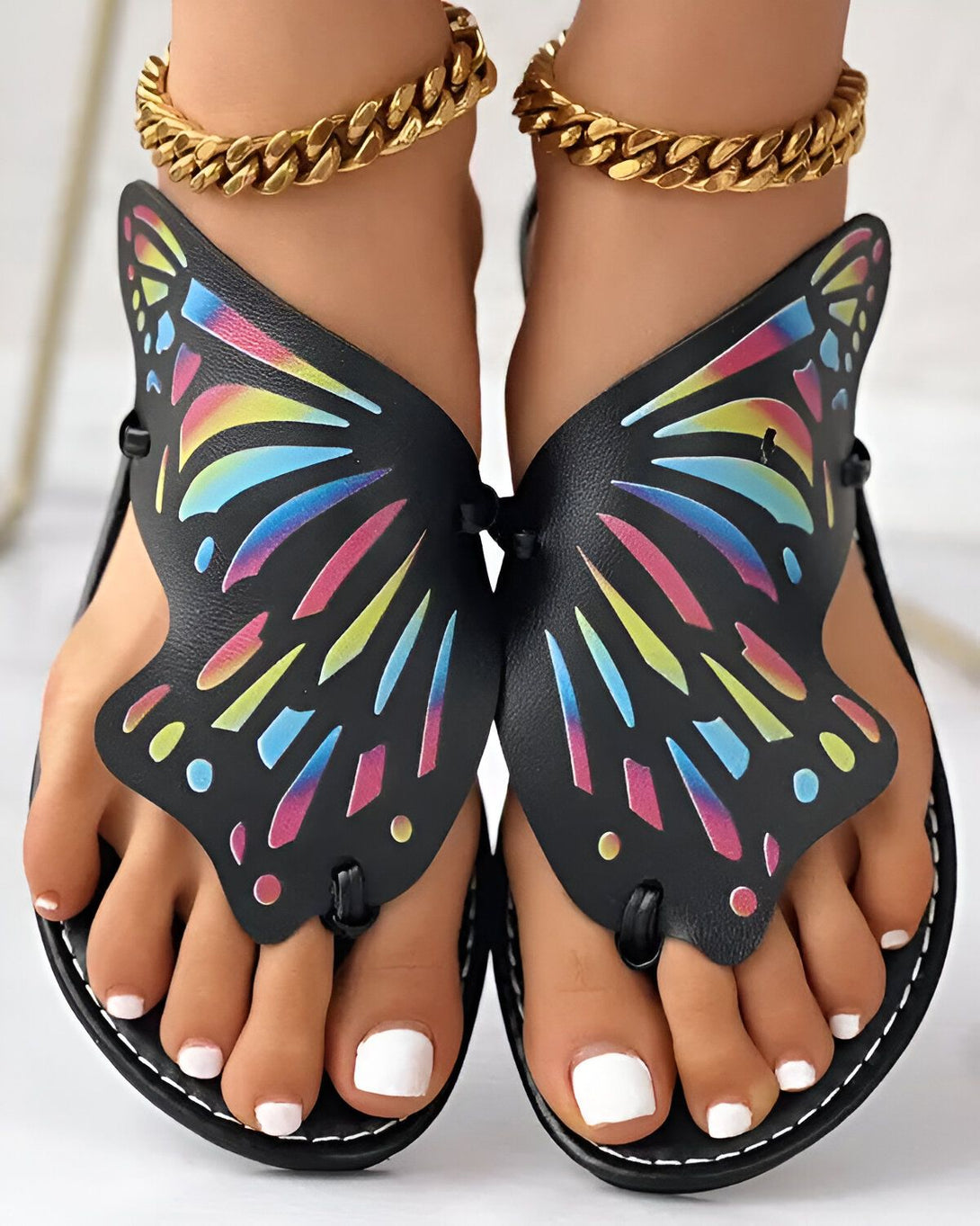 Une paire de sandales plates noires avec un motif de papillon coloré. Les sandales sont portées par une femme qui a les ongles des pieds peints en blanc. La femme porte également un bracelet de cheville doré.