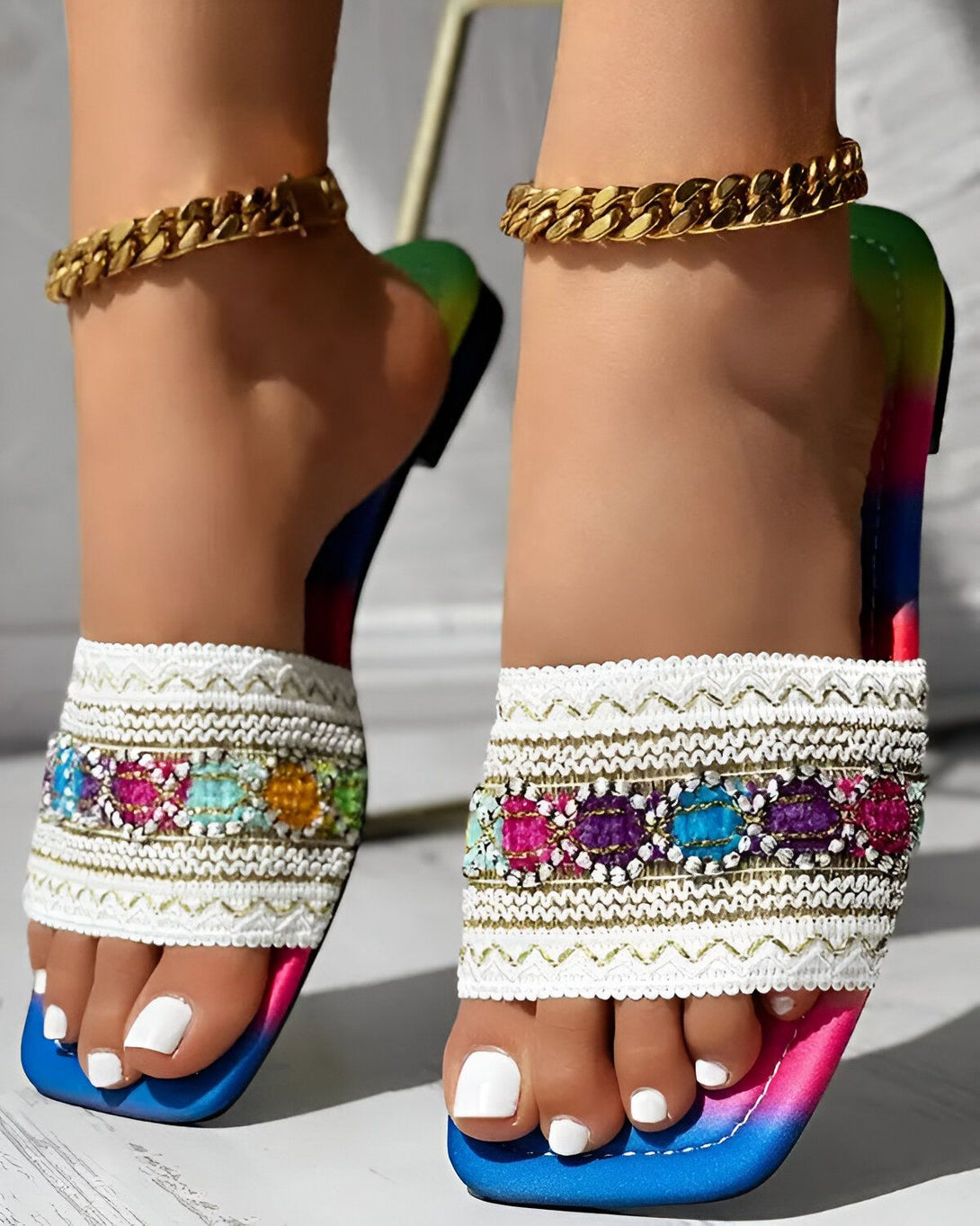 Une femme porte des sandales plates à enfiler. Les sandales sont blanches avec des broderies colorées et une semelle extérieure arc-en-ciel. La femme a les ongles des pieds peints en blanc et porte un bracelet de cheville en or.