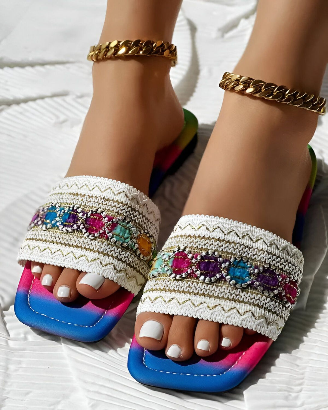 Une femme porte des sandales plates avec des perles et des pierres précieuses multicolores. Les sandales ont une semelle intérieure blanche et une semelle extérieure rose, bleue et violette. La femme a les ongles des pieds peints en blanc et porte un bracelet de cheville en or.