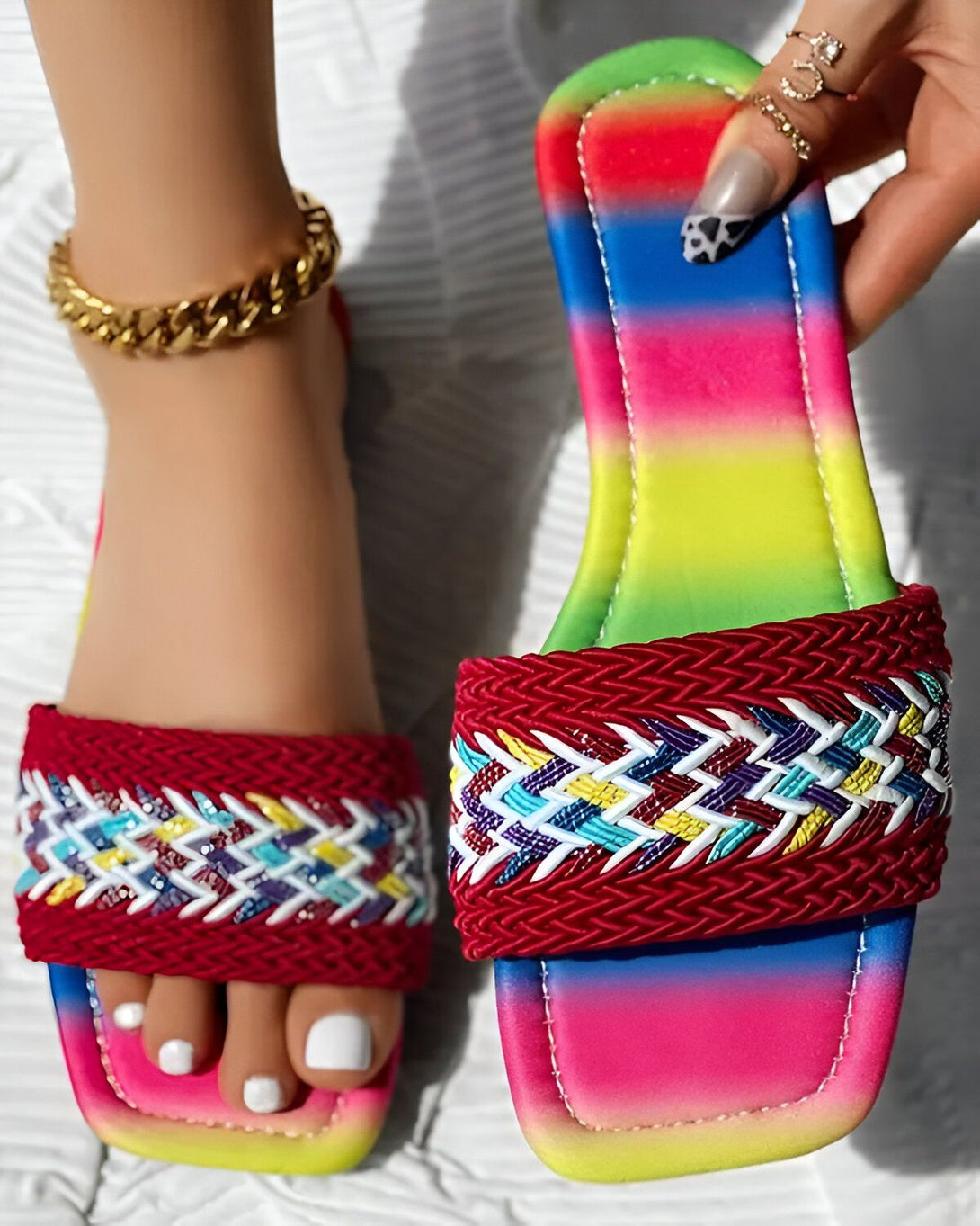 Une femme porte des sandales plates rouges et arc-en-ciel. Les sandales sont ornées de perles et de tresses colorées. La femme a les ongles peints en noir et blanc et porte un bracelet de cheville doré.