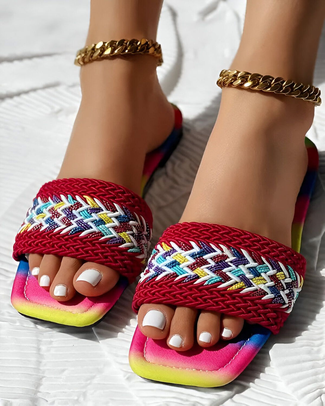 Une femme porte des sandales plates rouges, jaunes et vertes. Les sandales sont ornées d'un motif tressé et ont une semelle intérieure blanche. La femme a les ongles des pieds peints en blanc et porte un bracelet de cheville doré.