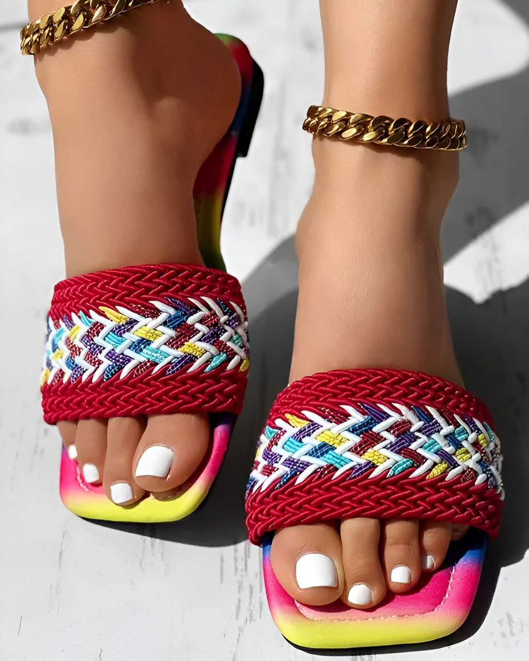 Une femme porte des sandales plates tissées à la main avec un motif coloré. Les sandales ont une semelle intérieure rembourrée et une semelle extérieure en caoutchouc. La femme a les ongles des pieds peints en blanc et porte un bracelet de cheville en or.