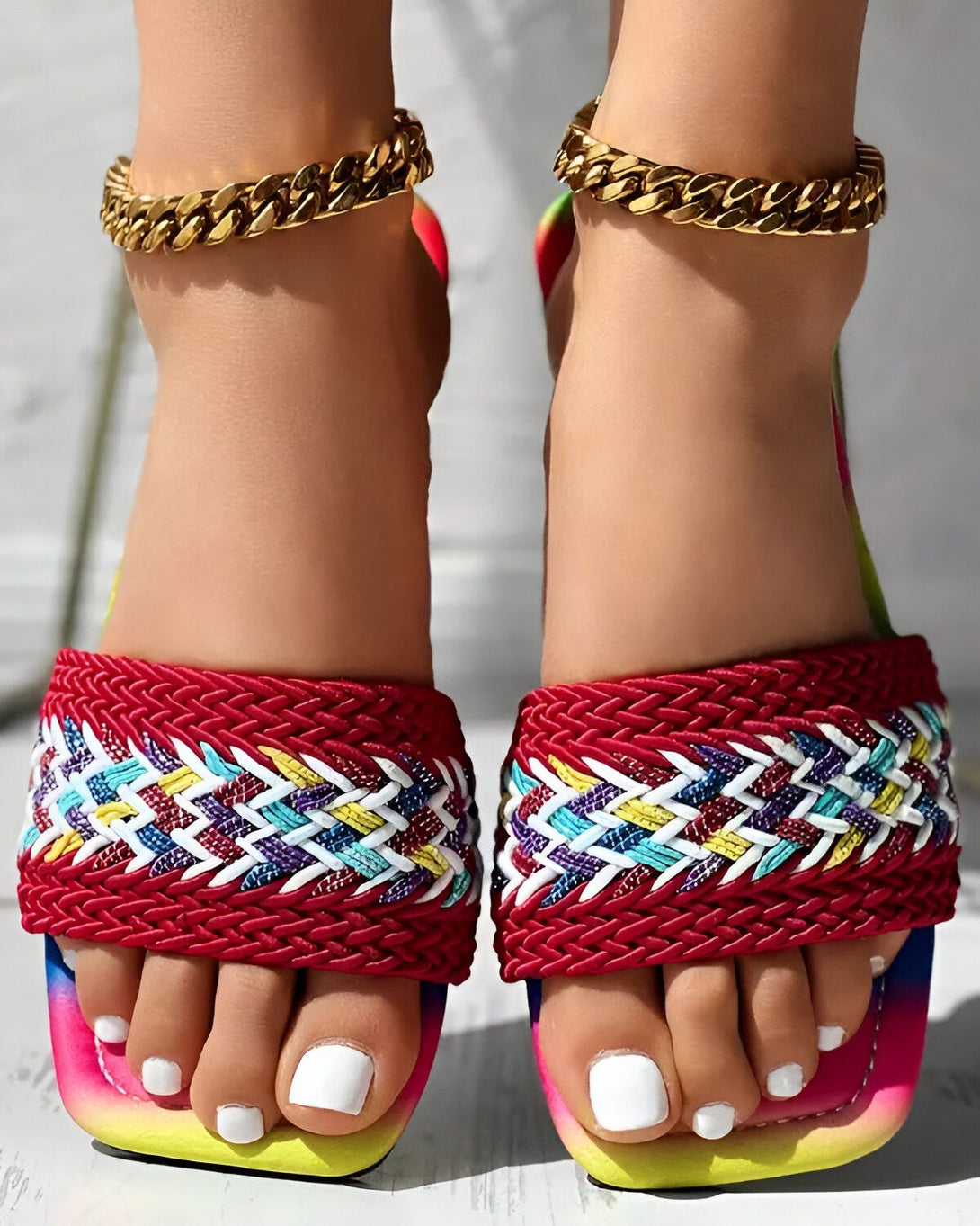 Une femme porte des sandales plates rouges, jaunes et bleues. Les sandales sont ornées d'un motif de chevrons et ont une semelle épaisse. La femme a les ongles des pieds peints en blanc et porte des bracelets de cheville en or.