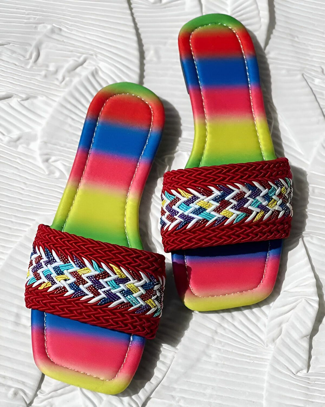 Une paire de sandales plates arc-en-ciel avec une bride tressée rouge vif. Les sandales sont posées sur un fond blanc.