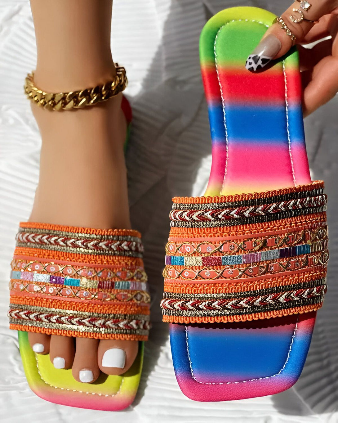 Une femme porte des sandales plates avec des perles et des broderies colorées. Les sandales ont une semelle intérieure orange et une semelle extérieure multicolore. La femme porte également un bracelet de cheville doré.